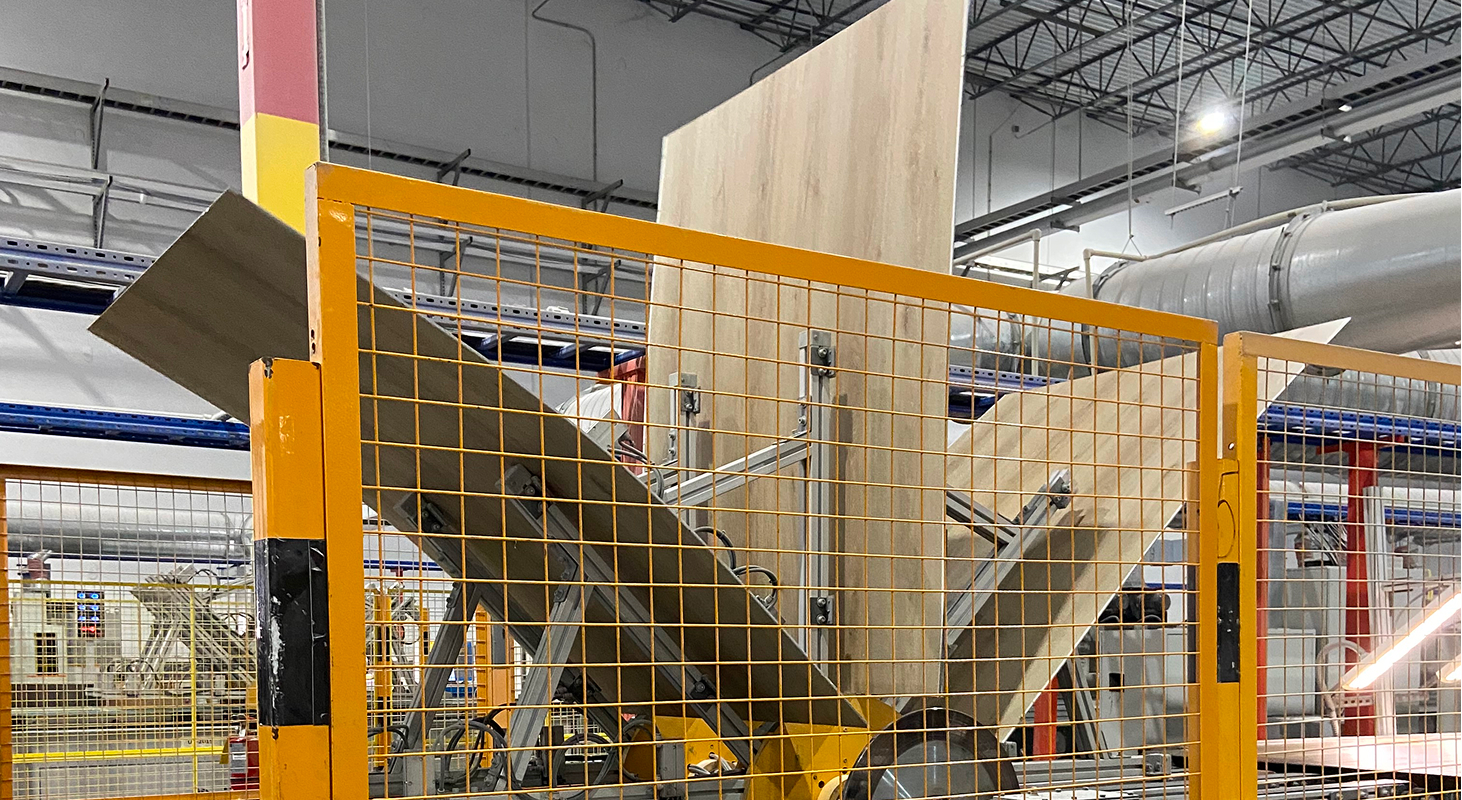 Large flooring panels moving through automated handling equipment inside an industrial manufacturing facility, with safety guards and production lines visible.